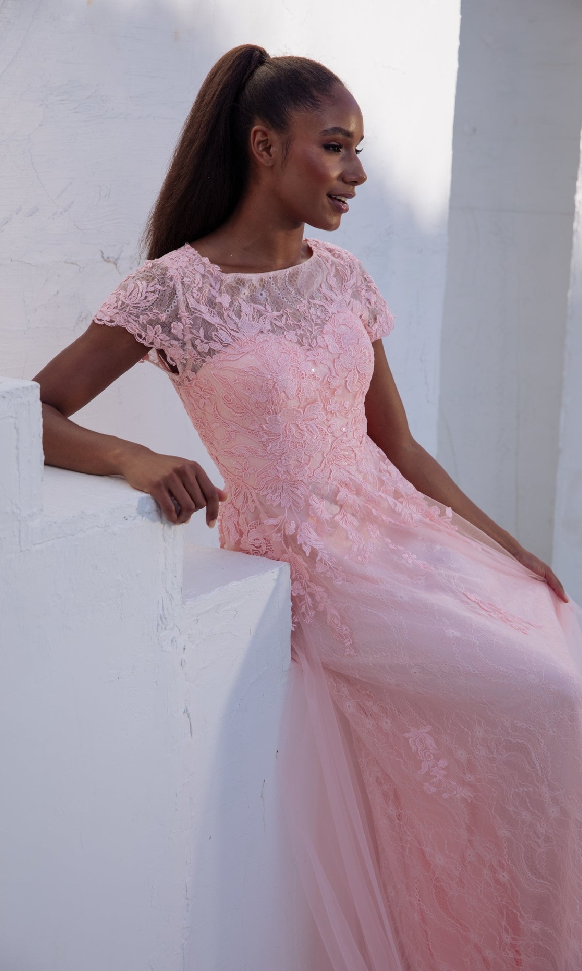 A woman in the Modest Short-Sleeve Prom Gown: Eureka EK104 sits on a white ledge, wearing a pink floral-lace applique evening dress with short sleeves and a sheer neckline, set against a white textured background.
