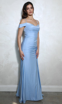 A woman stands against a light-colored wall wearing the Long Ruched Jersey Prom Dress: Eureka EK107, a floor-length, light blue off-the-shoulder gown with a fitted bodice and subtle shimmer.