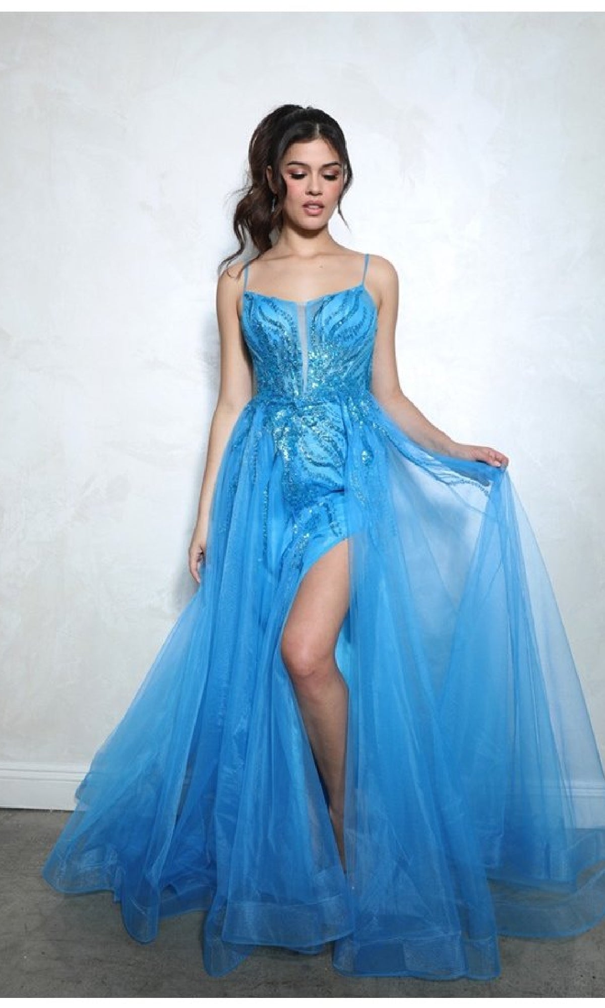 Woman wearing the Eureka EK112 Overskirt Long Sequin Prom Dress, a sleeveless bright blue gown with a sheer tulle overlay and high slit, standing against a plain light-colored wall.