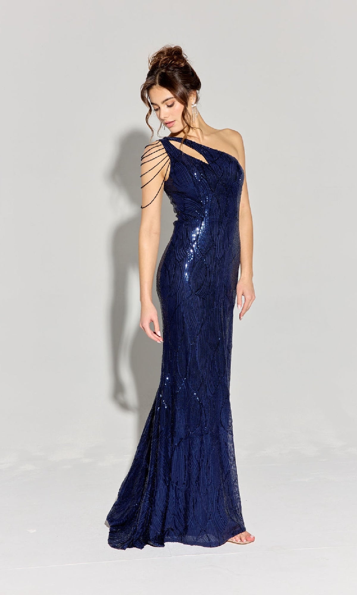 A woman wearing the Eureka EK113 One-Shoulder Long Sequin Prom Dress in navy stands on a plain light background, looking down to her left.