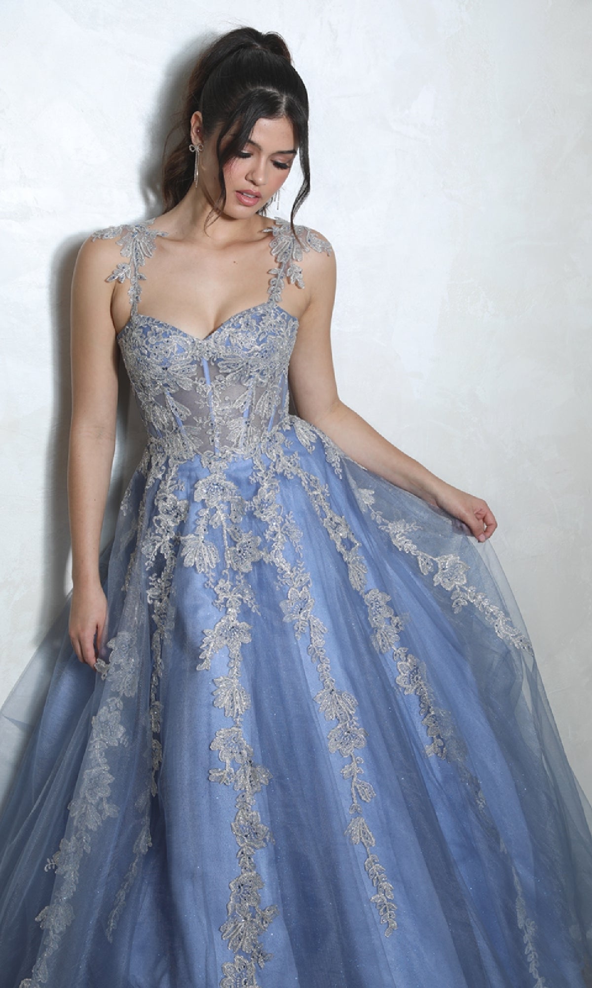 A woman wearing the Long Blue Sweetheart Prom Gown (Eureka EK115), featuring embroidered floral patterns and silver lace, stands against a light-colored textured wall, looking down.