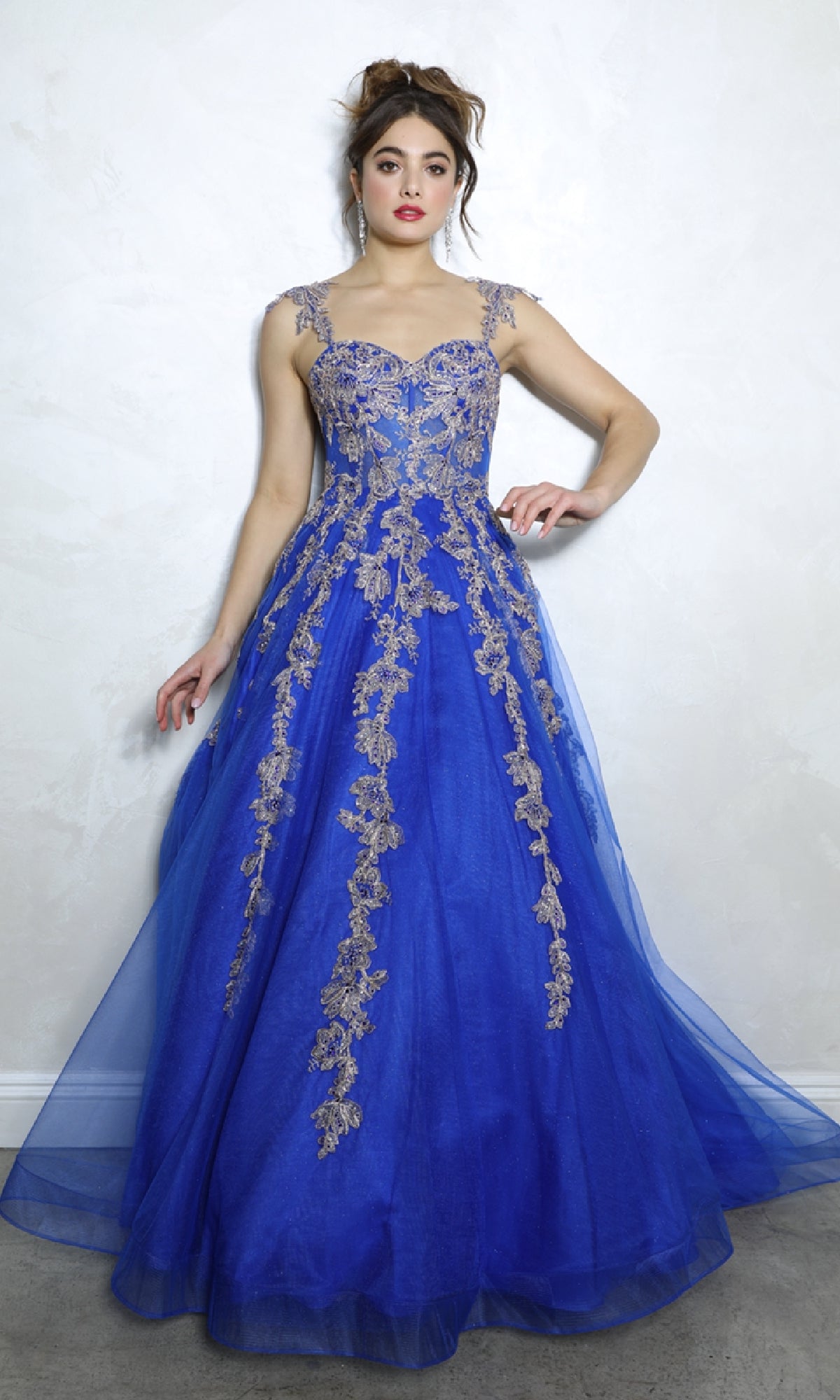 A woman stands against a light textured wall, wearing the Eureka EK115 Long Blue Sweetheart Prom Gown featuring embroidered florals, gold lace embellishments, and sheer straps.