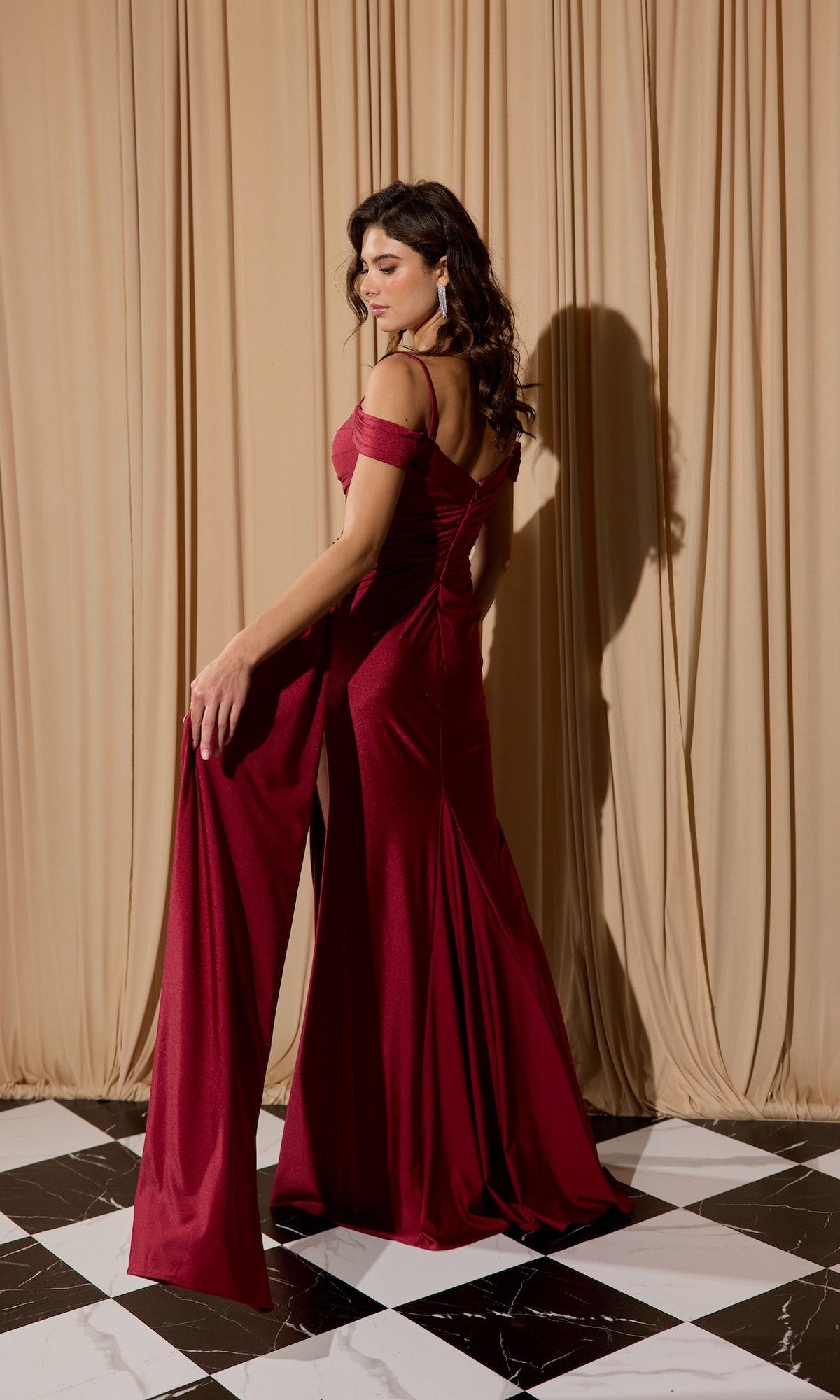 A woman wears the red Off-Shoulder Long Formal Dress: Eureka EK117 as she stands on a black and white checkered floor before beige curtains, glancing over her shoulder.