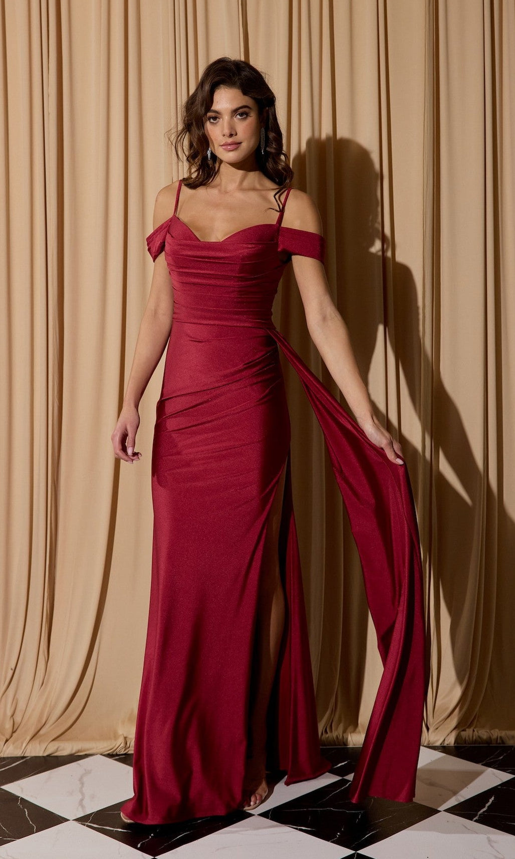 A woman poses gracefully before beige curtains, standing on a black and white tiled floor in the Eureka EK117—a burgundy off-shoulder long formal dress featuring a side slit.