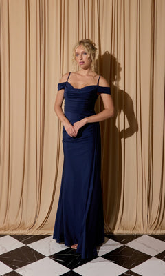A woman in a navy Eureka EK117 off-shoulder long formal dress stands on a black-and-white tiled floor before beige curtains.