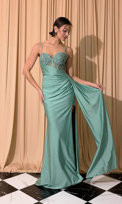 A woman wears the Long Prom Dress: Eureka EK166 in Agave, featuring a fitted, sheer-embroidered bodice and draped floor-length skirt, standing on black and white tiles before beige curtains.
