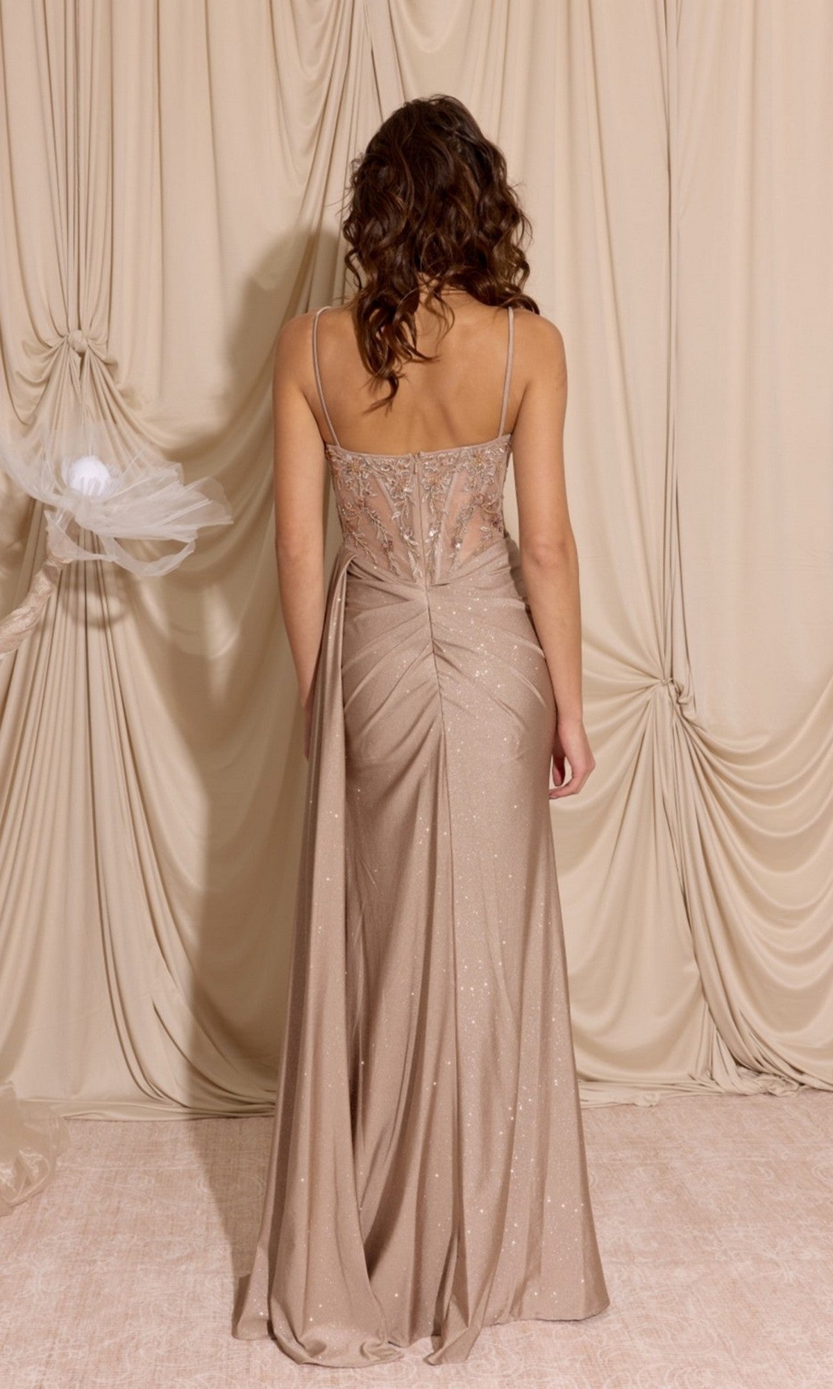 A woman with wavy hair faces away, wearing the Eureka EK166 long prom dress in beige, featuring a sheer-embroidered bodice and sparkling details, standing before draped beige fabric.