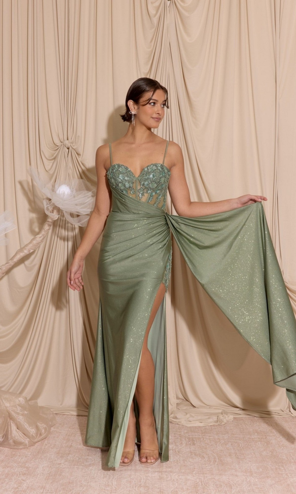 Wearing the Eureka EK166 long prom dress in cactus green with a thigh-high slit and draped detailing, a woman stands before beige curtains.