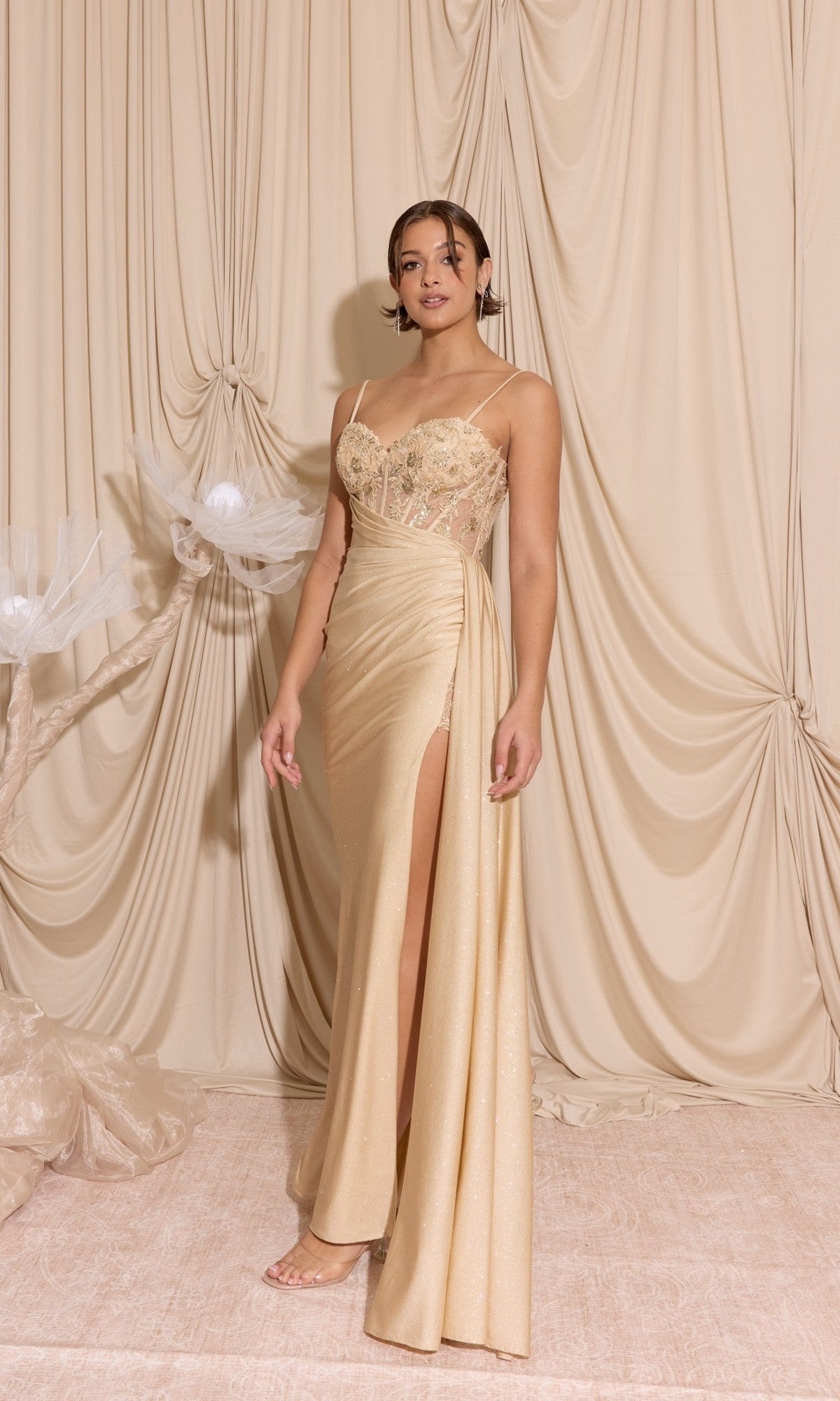 A woman poses before draped beige fabric, wearing the Eureka EK166 Long Prom Dress in Champagne, featuring a sheer-embroidered bodice, thin straps, delicate beading, and a high slit.