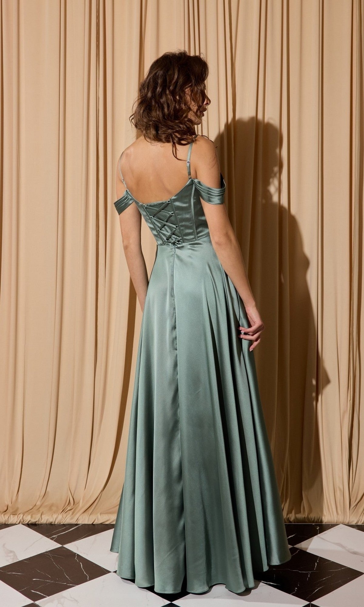 A woman stands facing away in front of beige curtains, wearing the Eureka EK176 long prom dress—a floor-length satin gown in sage green with an off-shoulder bodice and flowing skirt.