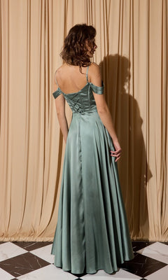 A woman stands facing away in front of beige curtains, wearing the Eureka EK176 long prom dress—a floor-length satin gown in sage green with an off-shoulder bodice and flowing skirt.