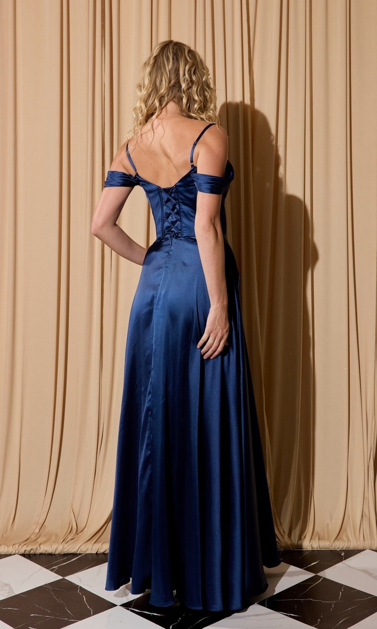 A woman with curly blonde hair stands with her back to the camera on a checkered floor, wearing the Eureka EK176 long satin prom dress in deep blue, set against beige drapes.