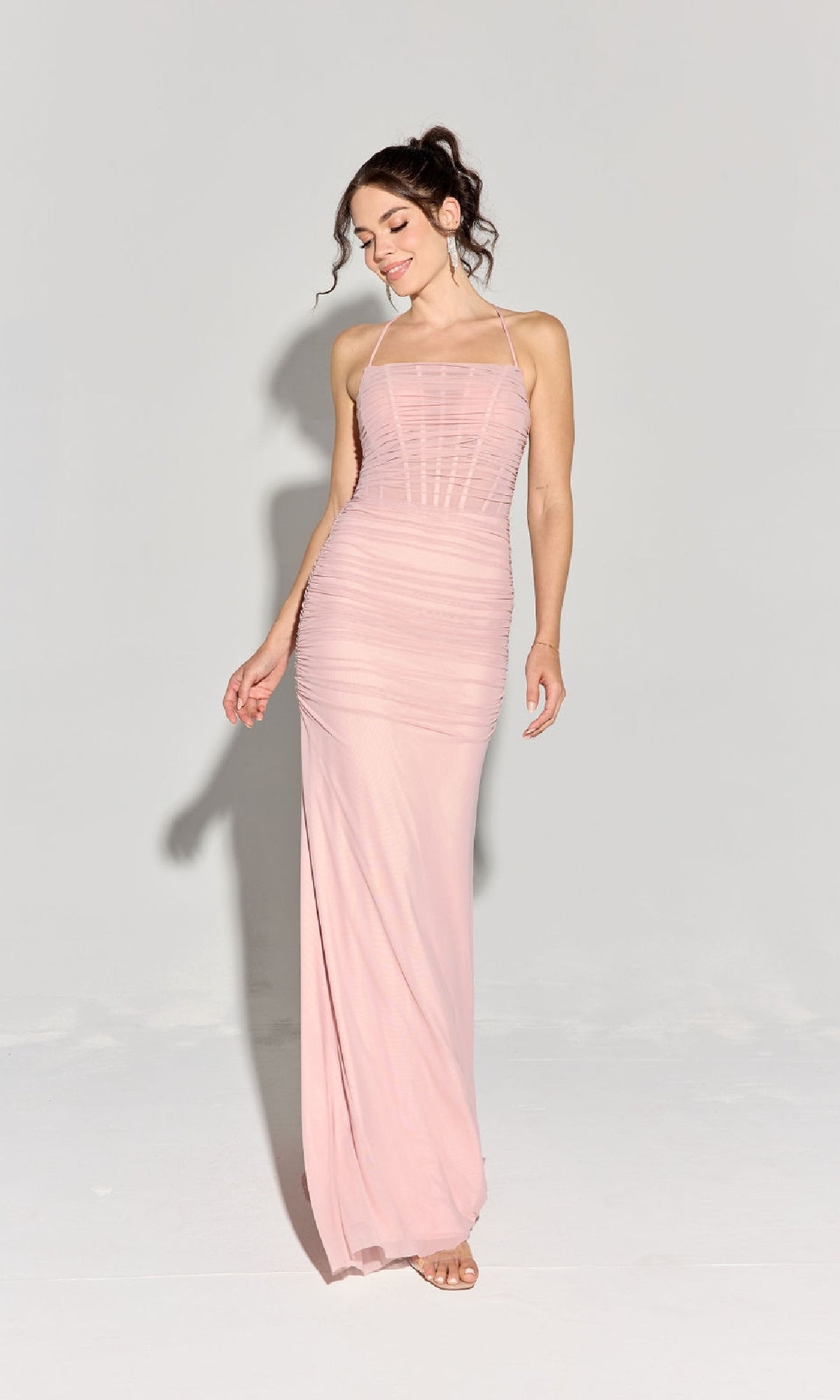 A woman wearing the Strappy-Back Slinky Long Prom Dress: Eureka EK177 in light pink stands against a plain background, looking down and smiling.