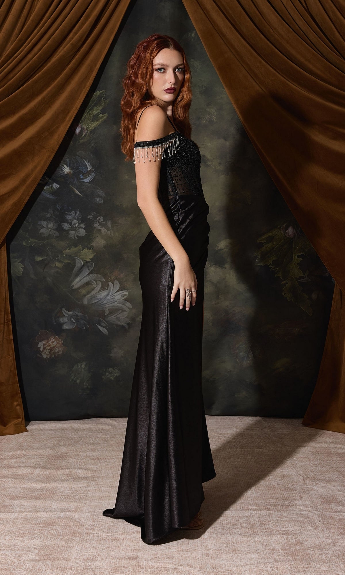 A person wears a black Beaded-Corset Long Satin Prom Dress: Eureka EK179, featuring a beaded corset bodice and off-the-shoulder straps with dangling crystal fringe.