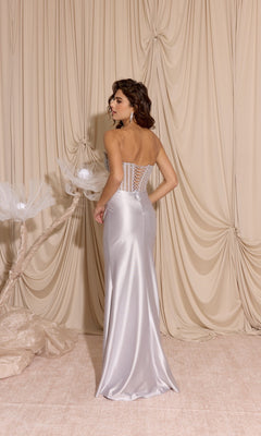 A woman in the Eureka EK183 strapless tight satin prom dress with a corset back stands before beige draped curtains and decorative white flowers.