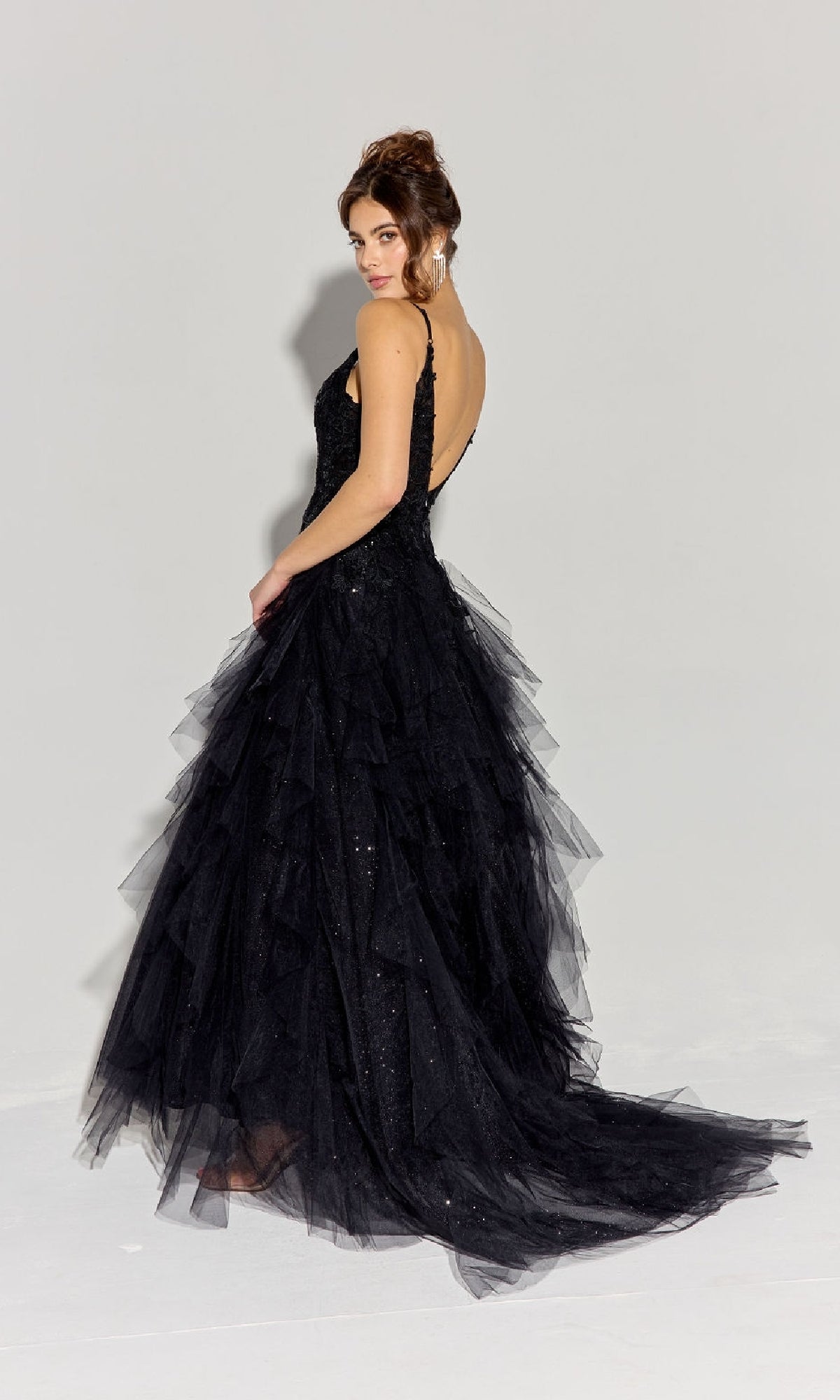 Woman wearing the Ruffled Long Glitter Prom Ball Gown: Eureka EK188—a sleeveless, floor-length black tulle dress with an open back and layered skirt—standing and looking over her shoulder against a plain light background.