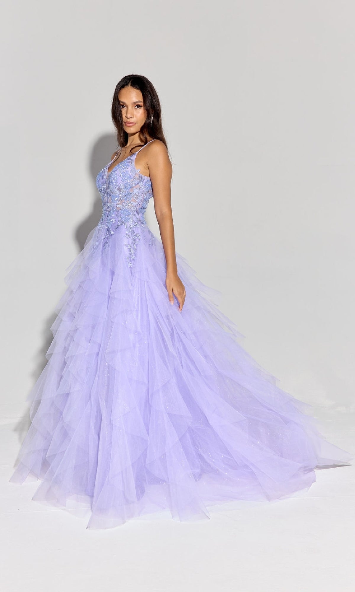 A woman in the Ruffled Long Glitter Prom Ball Gown: Eureka EK188, a light purple, floor-length tulle dress with floral embroidery, stands against a plain background, showcasing the elegance and sparkle of this glamorous evening gown.