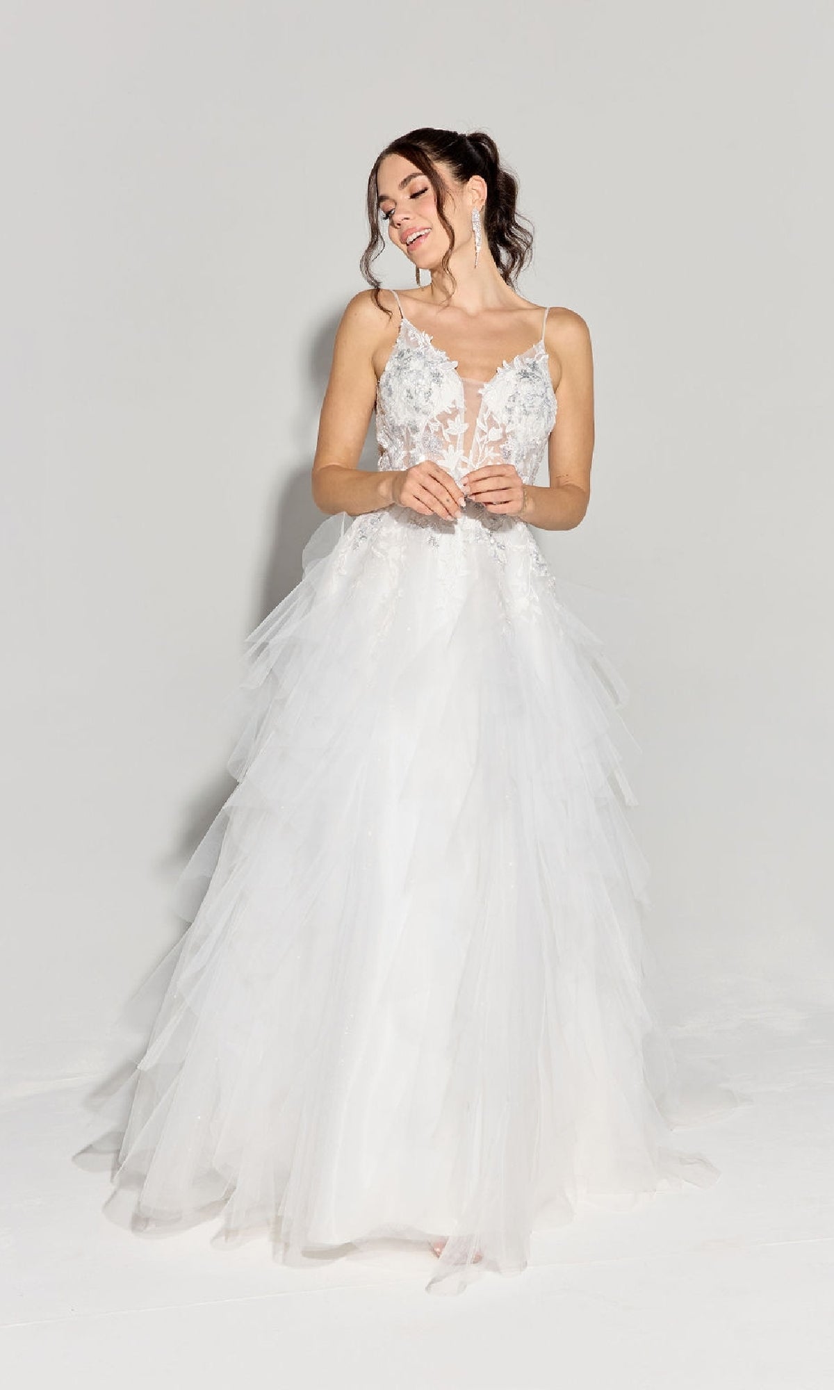 A woman in the Ruffled Long Glitter Prom Ball Gown, Eureka EK188, featuring a white layered tulle skirt and lace floral bodice, stands against a plain light background, looking left and smiling.