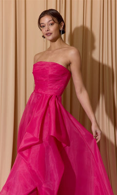 A woman in the Long Prom Dress: Eureka EK206, a strapless bright pink organza gown with a layered skirt, stands before beige curtains—perfect for anyone seeking a standout ball gown look.