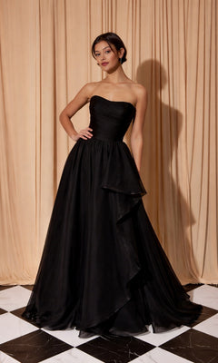 A woman in the Eureka EK206 long prom dress in black stands on a black and white checkered floor before beige curtains, her strapless organza gown radiating sophisticated elegance.