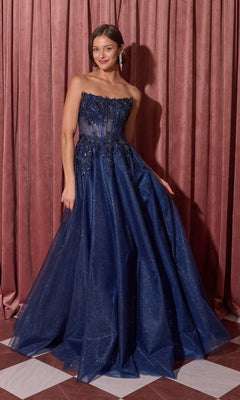 A woman in the Eureka EK208 long prom dress in navy, featuring a strapless beaded floral applique bodice and glitter tulle skirt, stands on a checkered floor before pink curtains, radiating elegance.