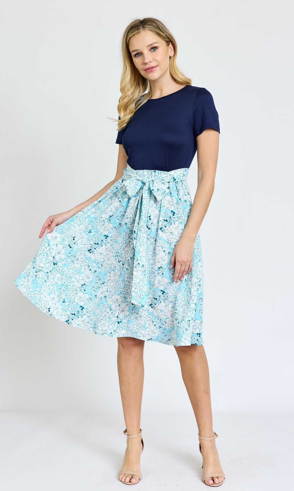 A woman stands against a white background wearing the Knee-Length Floral Casual Dress with Pockets and beige heels, holding out one side of the skirt.