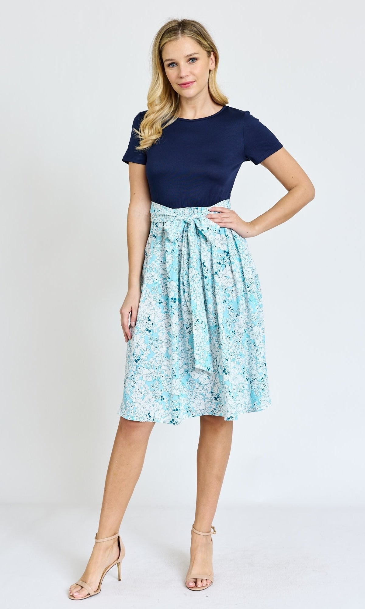 A woman stands against a plain background, wearing a knee-length floral casual dress with pockets, paired with nude heels.