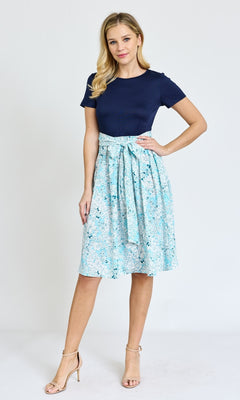 A woman stands against a plain background, wearing a knee-length floral casual dress with pockets, paired with nude heels.