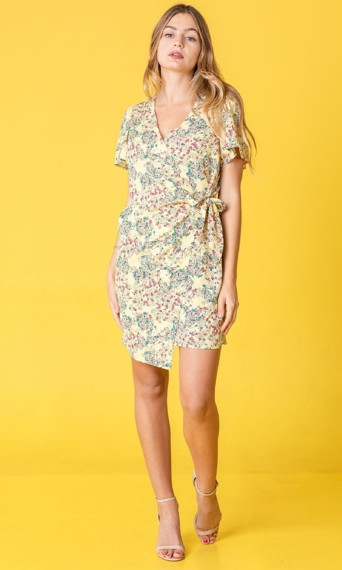 A woman stands against a yellow background wearing the Floral Print Casual Short Wrap Dress with short sleeves, a v-neckline, and beige high-heeled sandals.
