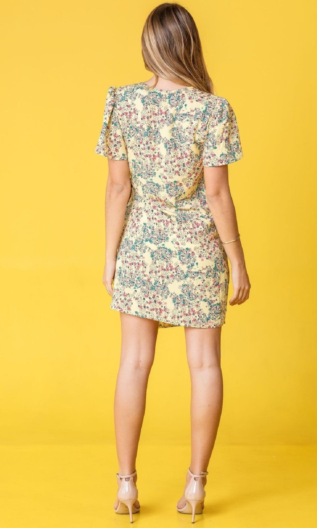 A woman stands facing away, wearing the Floral Print Casual Short Wrap Dress and nude heels, set against a solid yellow background.