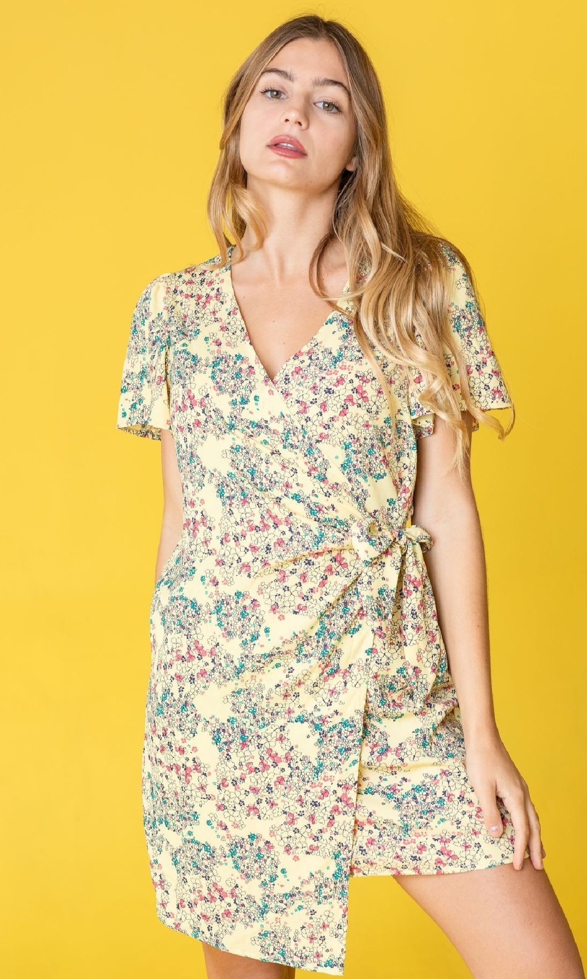 A woman wearing the Floral Print Casual Short Wrap Dress with a v-neckline stands against a yellow background, facing the camera with one hand by her side.