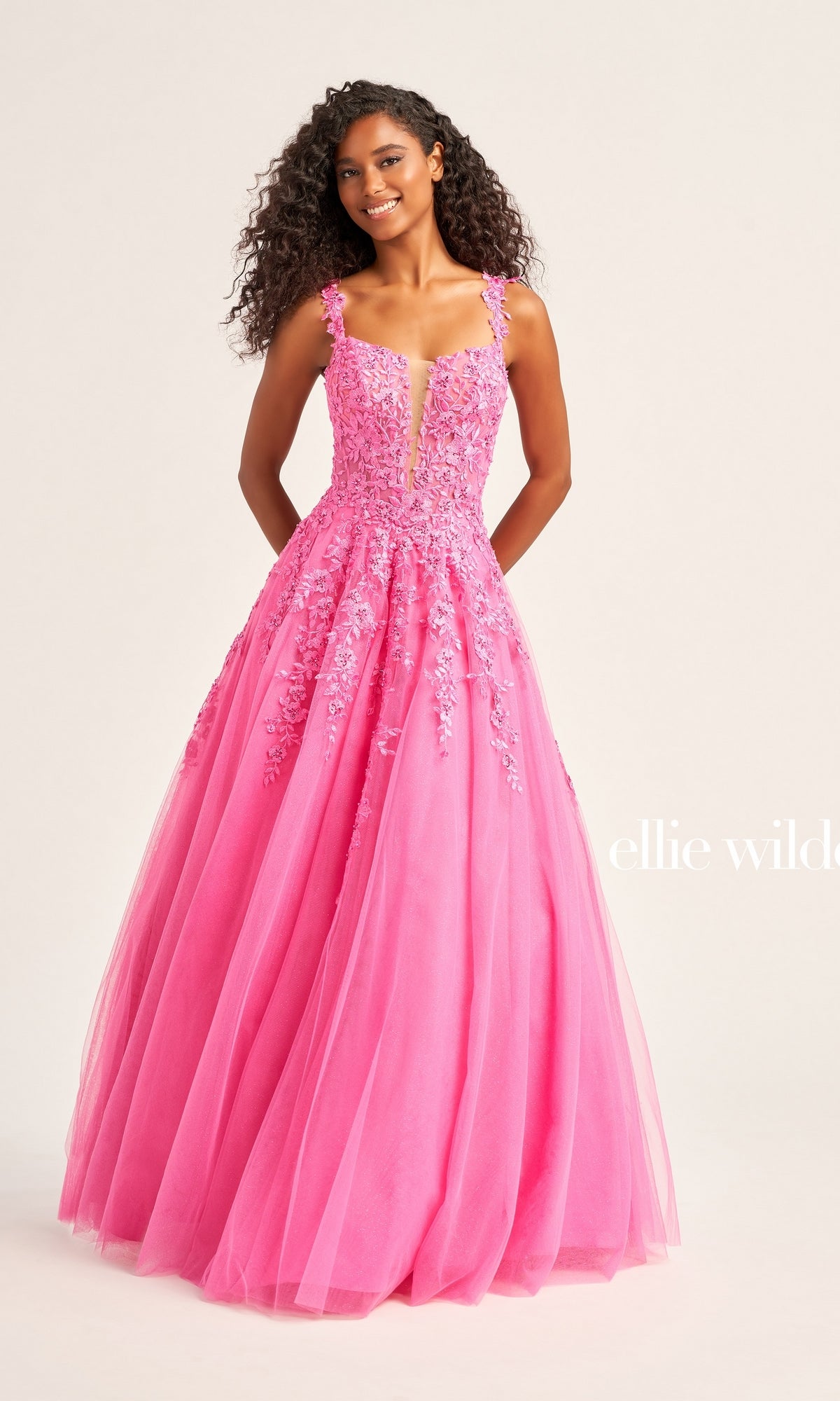 A woman in the Ellie Wilde EW120014 long glitter prom ball gown with floral appliqué details and a tulle skirt stands against a plain light background.