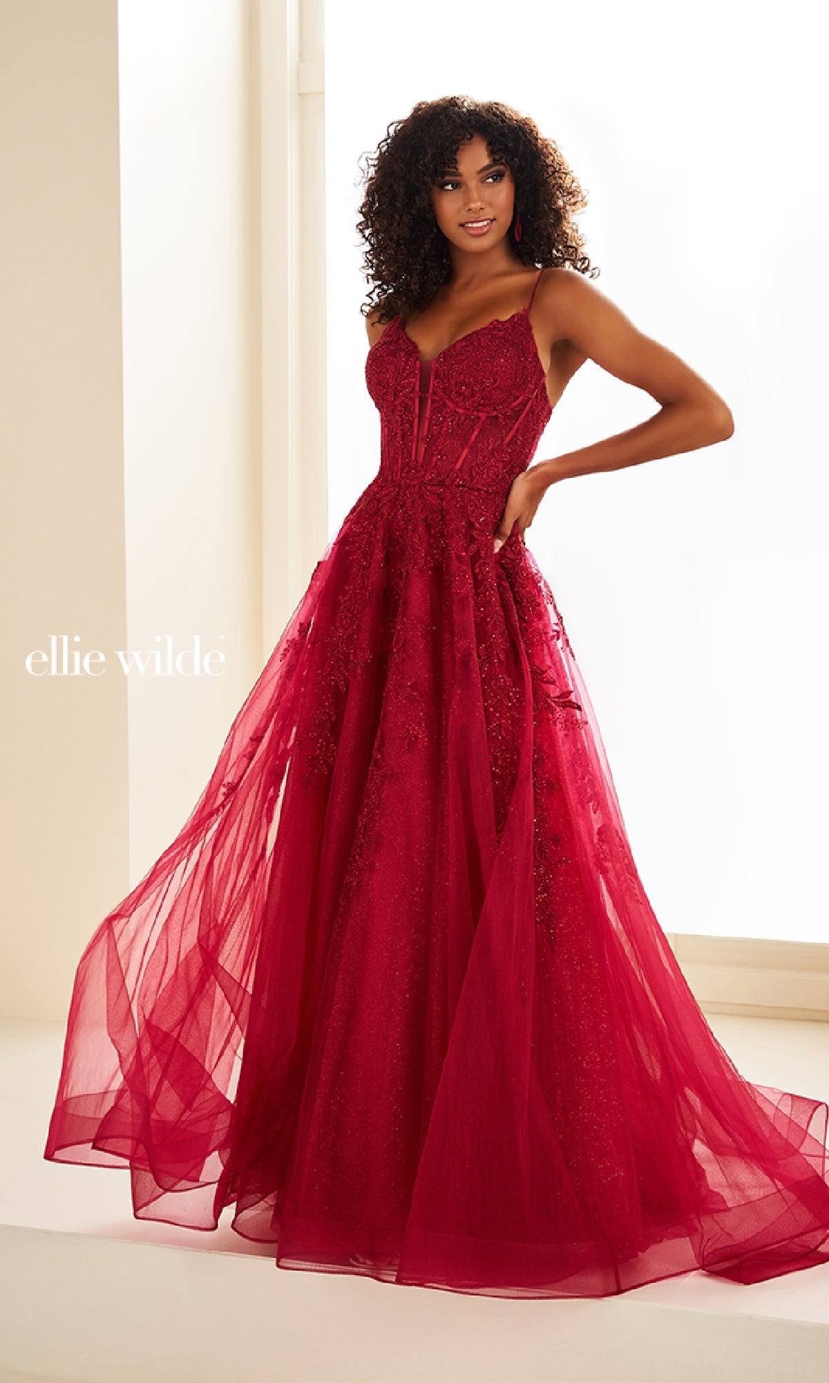 A woman stands in a brightly lit room wearing the Sheer-Corset Long Lace Prom Ball Gown EW34036, a fitted, floor-length red dress with lace and tulle detailing.