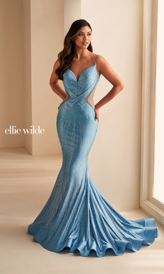 A woman stands near a window wearing the Sheer-Side Ellie Wilde Designer Prom Dress EW35001, a shimmering light blue, form-fitting mermaid gown with beading, thin straps, and sheer side cutouts.