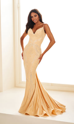 A woman stands by a bright window wearing the Ellie Wilde Long Designer Prom Dress EW35002—a form-fitting, light gold mermaid gown with thin straps and subtle embellishments.