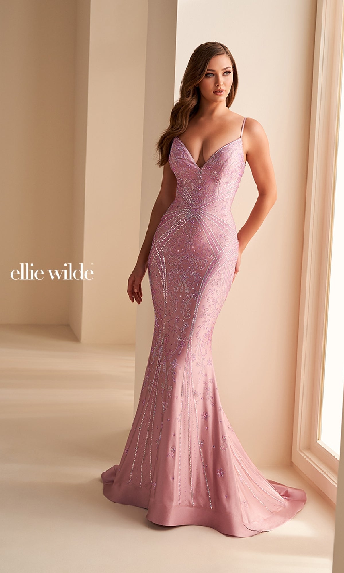 A woman wears the Ellie Wilde Long Designer Prom Dress EW35002, a fitted lavender gown with beading and thin straps, standing by a softly lit window. The 