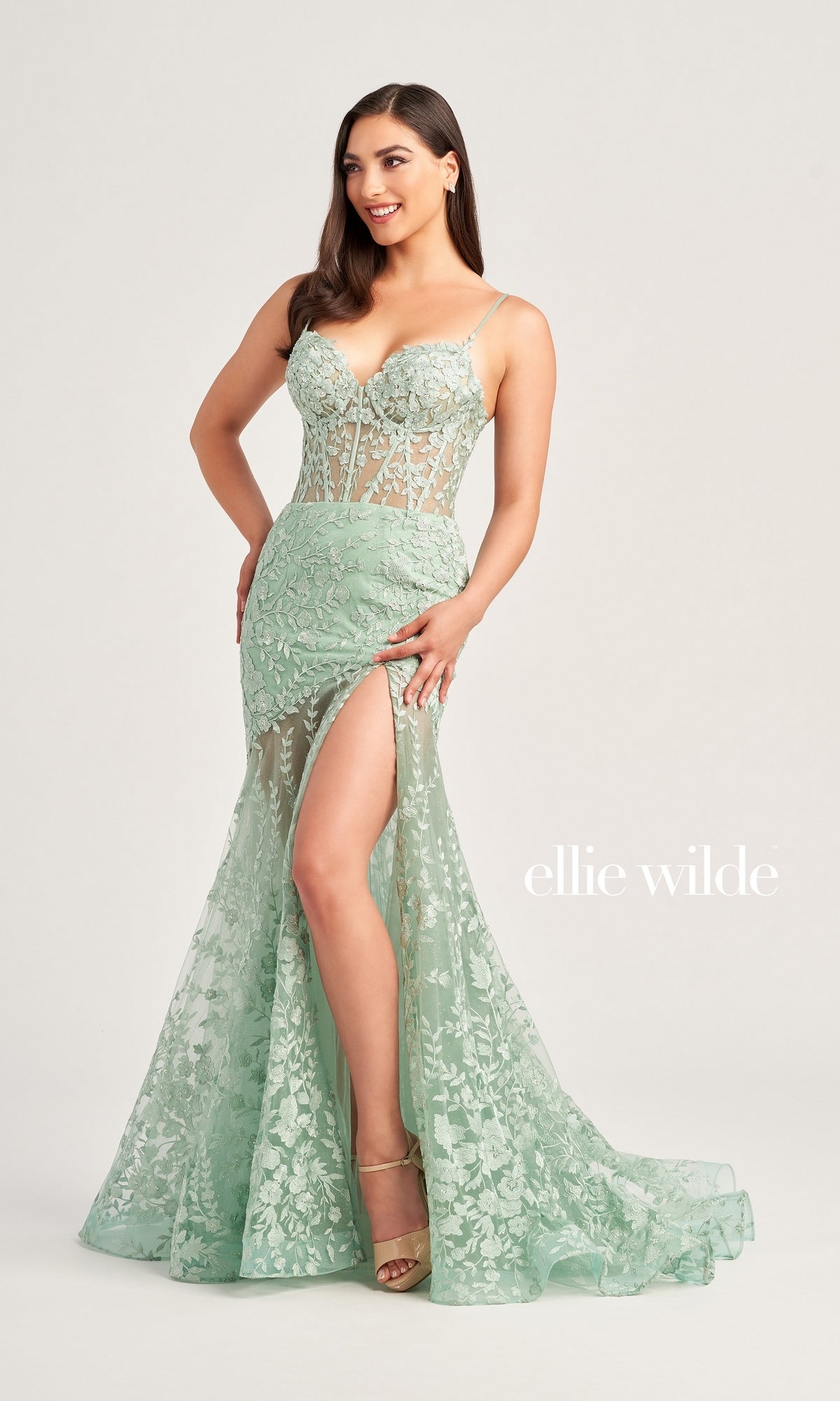 A woman models the Ellie Wilde Long Lace Mermaid Prom Dress EW35005 in light green, featuring a floor-length lace design with a high slit and spaghetti straps, against a plain backdrop. The text 