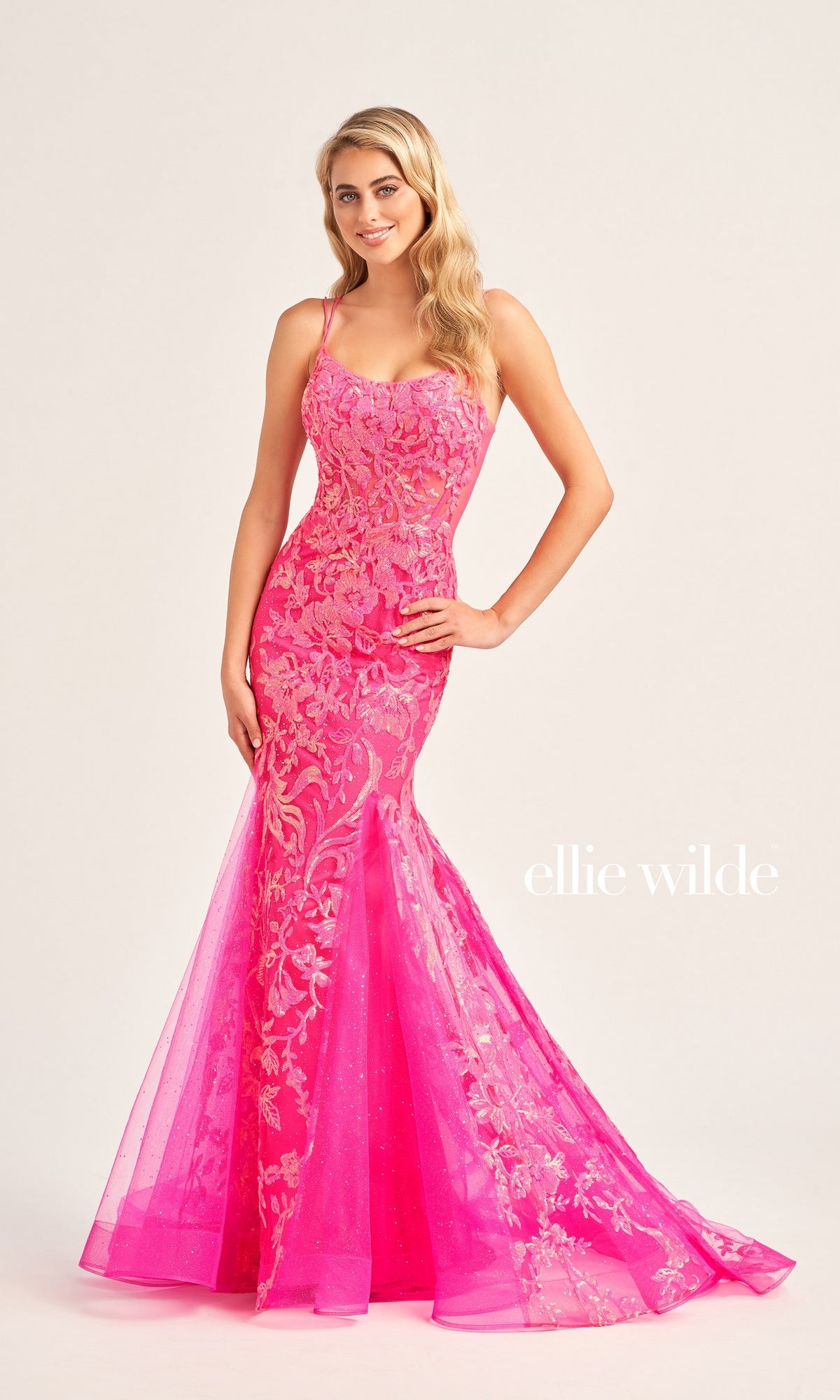 A woman poses with one hand on her hip, wearing the Ellie Wilde Sequin Long Mermaid Prom Dress EW35008—a fitted, bright pink floral lace sequin gown with thin straps—against a plain background. 