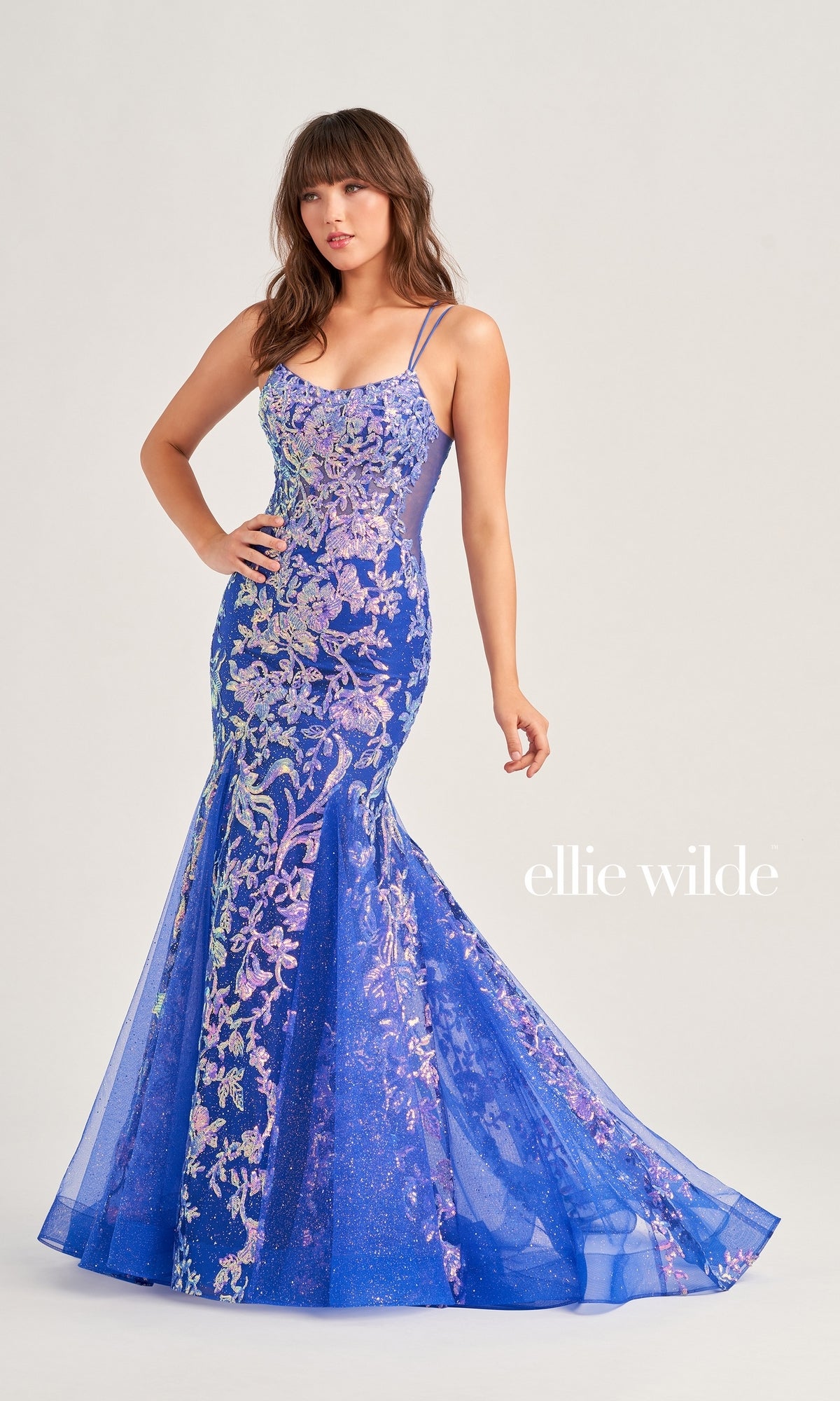 A woman wears the Ellie Wilde Sequin Long Mermaid Prom Dress EW35008, a fitted blue mermaid gown with floral embroidery and spaghetti straps, standing against a plain background. 