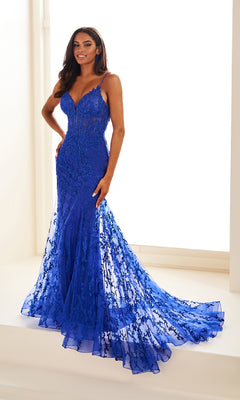 A woman stands indoors wearing the sleeveless, form-fitting Ellie Wilde Beaded-Tulle Long Prom Dress EW35010 in royal blue, featuring beaded embroidery, a mermaid silhouette, and a long train.