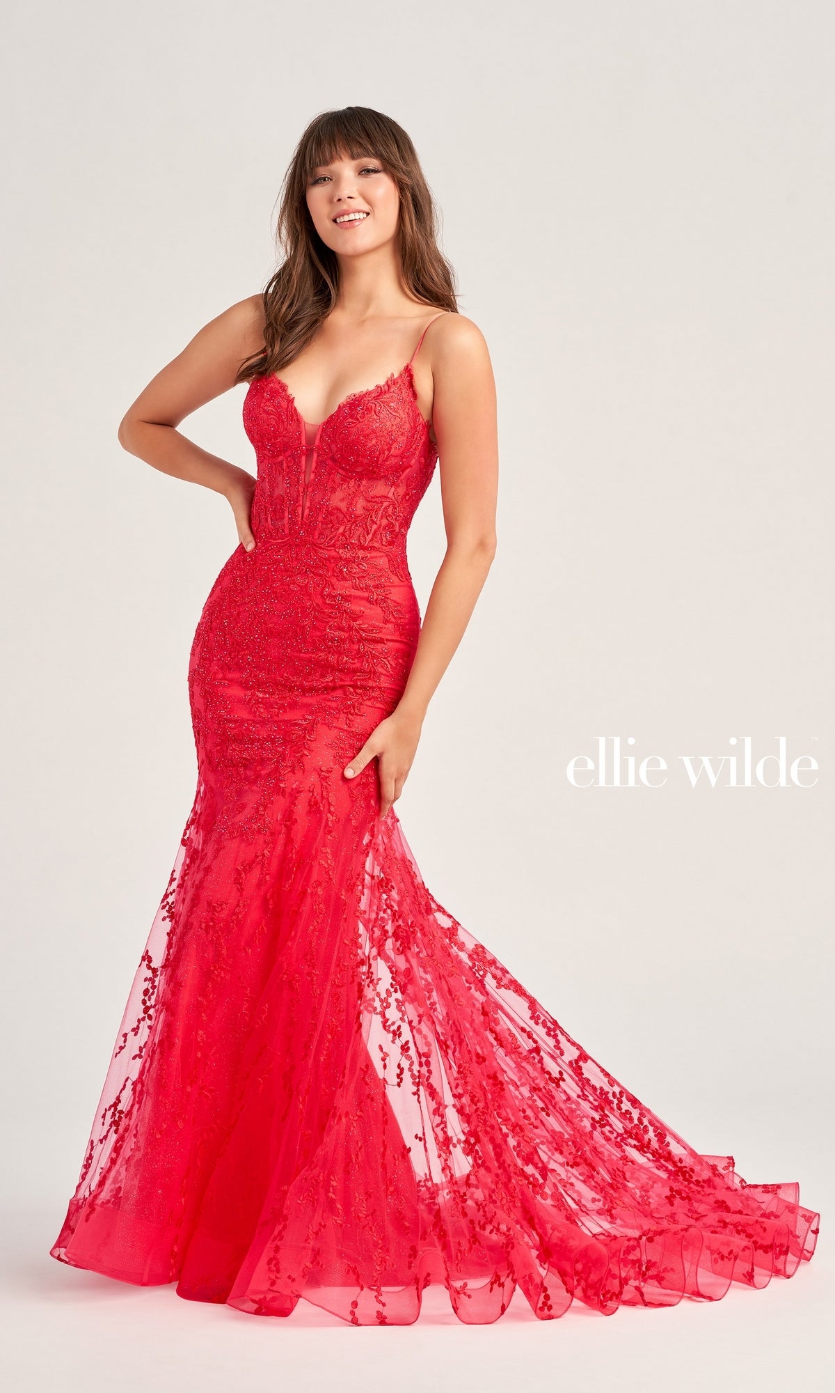 A woman wears the Ellie Wilde Beaded-Tulle Long Prom Dress EW35010—a fitted, floor-length red gown with thin straps, beaded embroidery, and a flared skirt—posing against a plain background with 