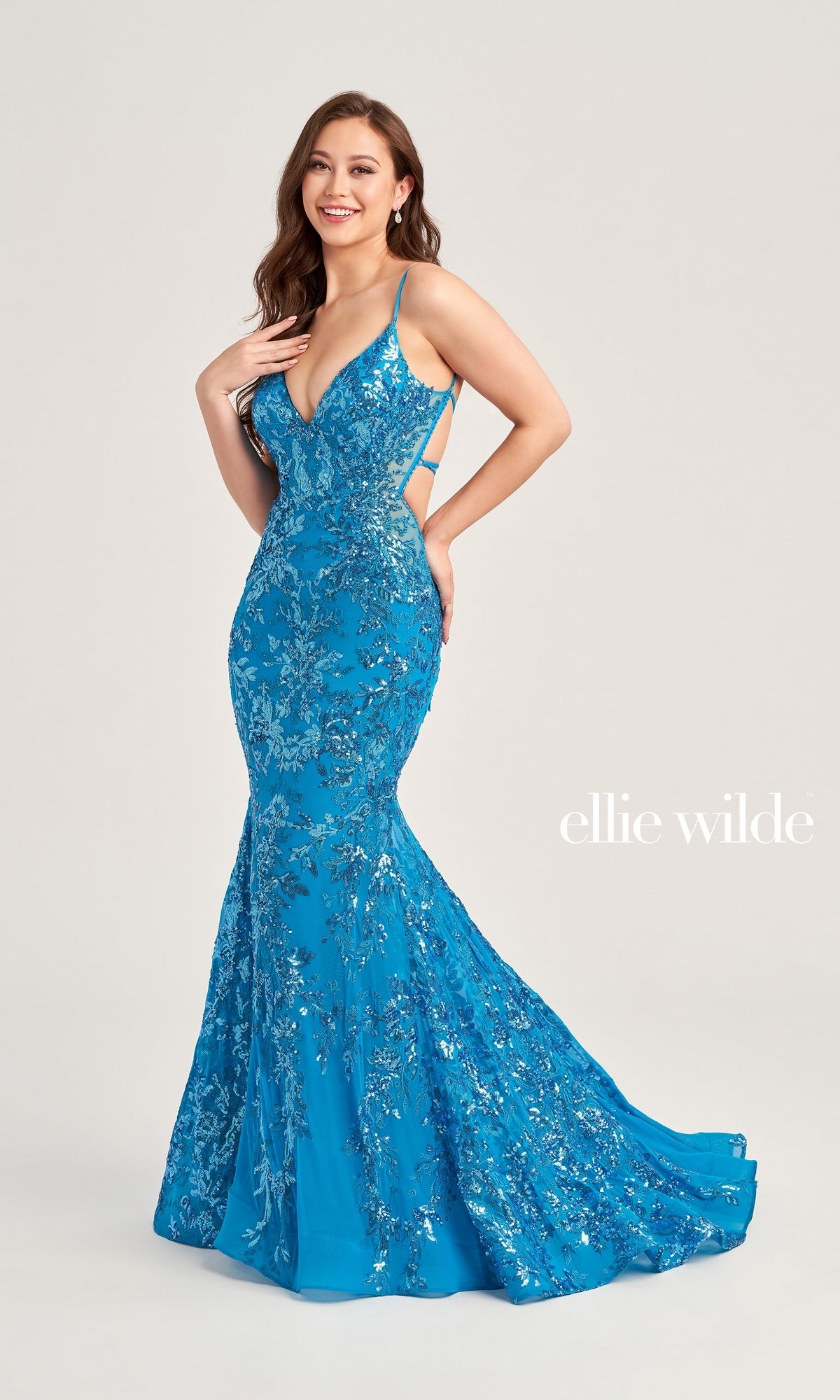 A woman poses in front of a plain background wearing the blue, sequin-covered Ellie Wilde Long Tulle Designer Prom Dress EW35011 with thin straps. The words 