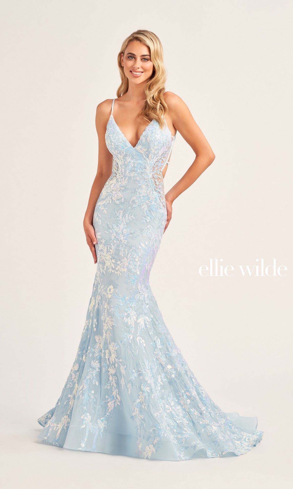 A woman poses in the Ellie Wilde Long Tulle Designer Prom Dress EW35011, featuring light blue tulle, sequin floral embroidery, a fitted bodice, and flared hem that highlight its elegant style against a plain background.