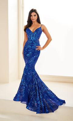 A woman stands indoors wearing the Ellie Wilde Long Tulle Designer Prom Dress EW35011, a fitted, floor-length gown with thin straps, flared hem, blue sequins, and elegant floral accents.
