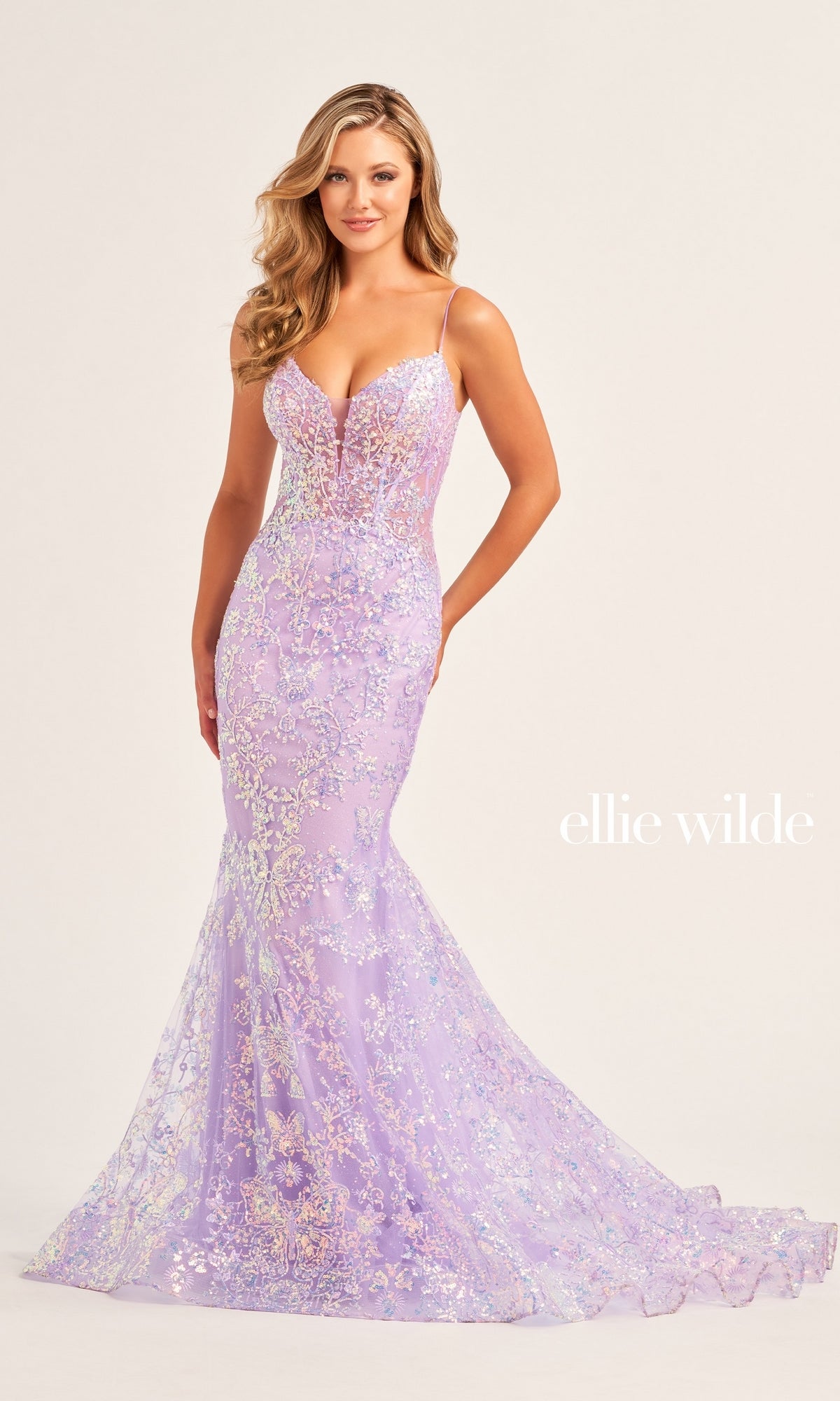 A woman poses in the Ellie Wilde Designer Glitter Prom Dress EW35013, featuring a fitted lavender sequin mermaid silhouette, sheer bodice, thin straps, and a flared train. The text 