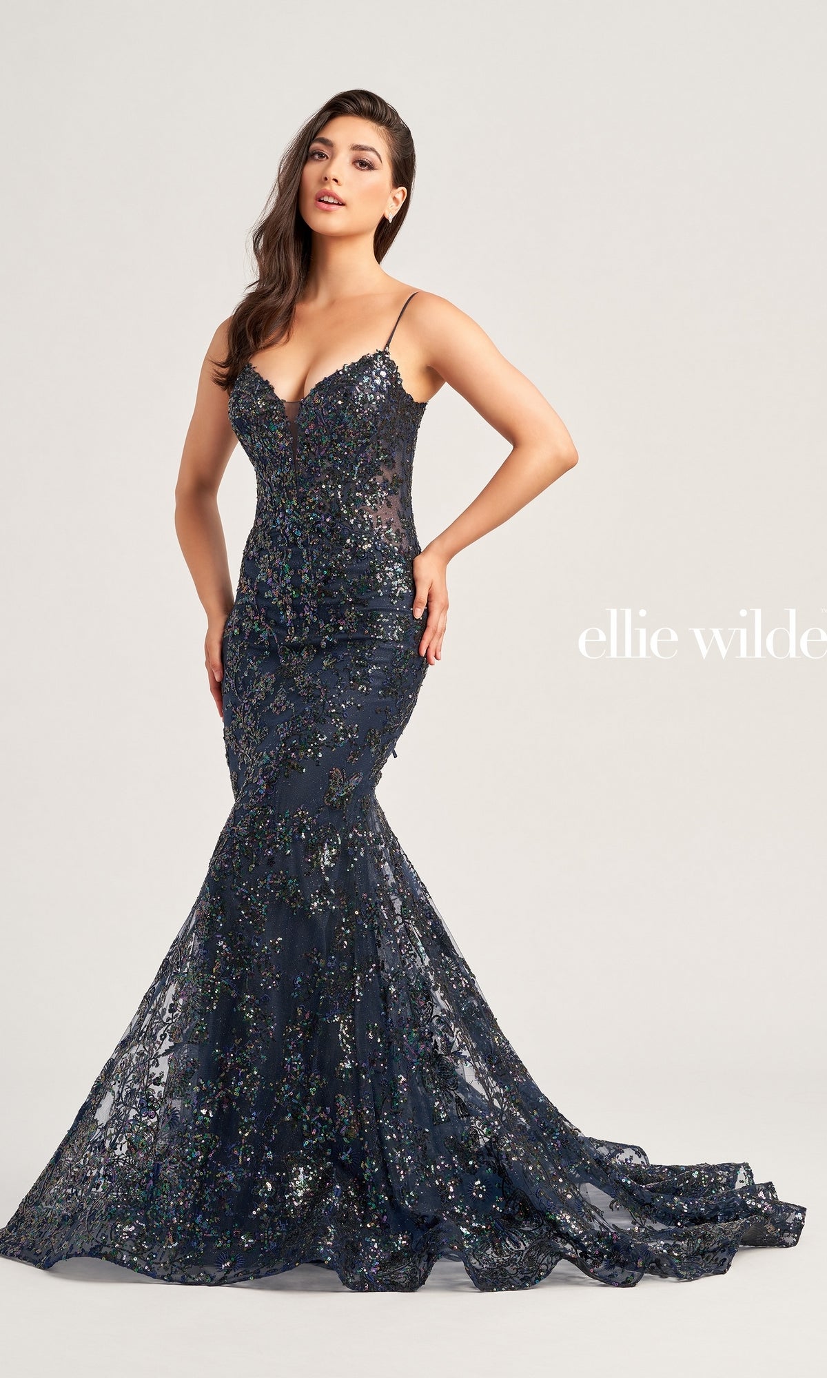 A woman poses against a plain light background in the Ellie Wilde Designer Glitter Prom Dress EW35013, a navy blue, strapless mermaid gown with sequin embellishments. The text 