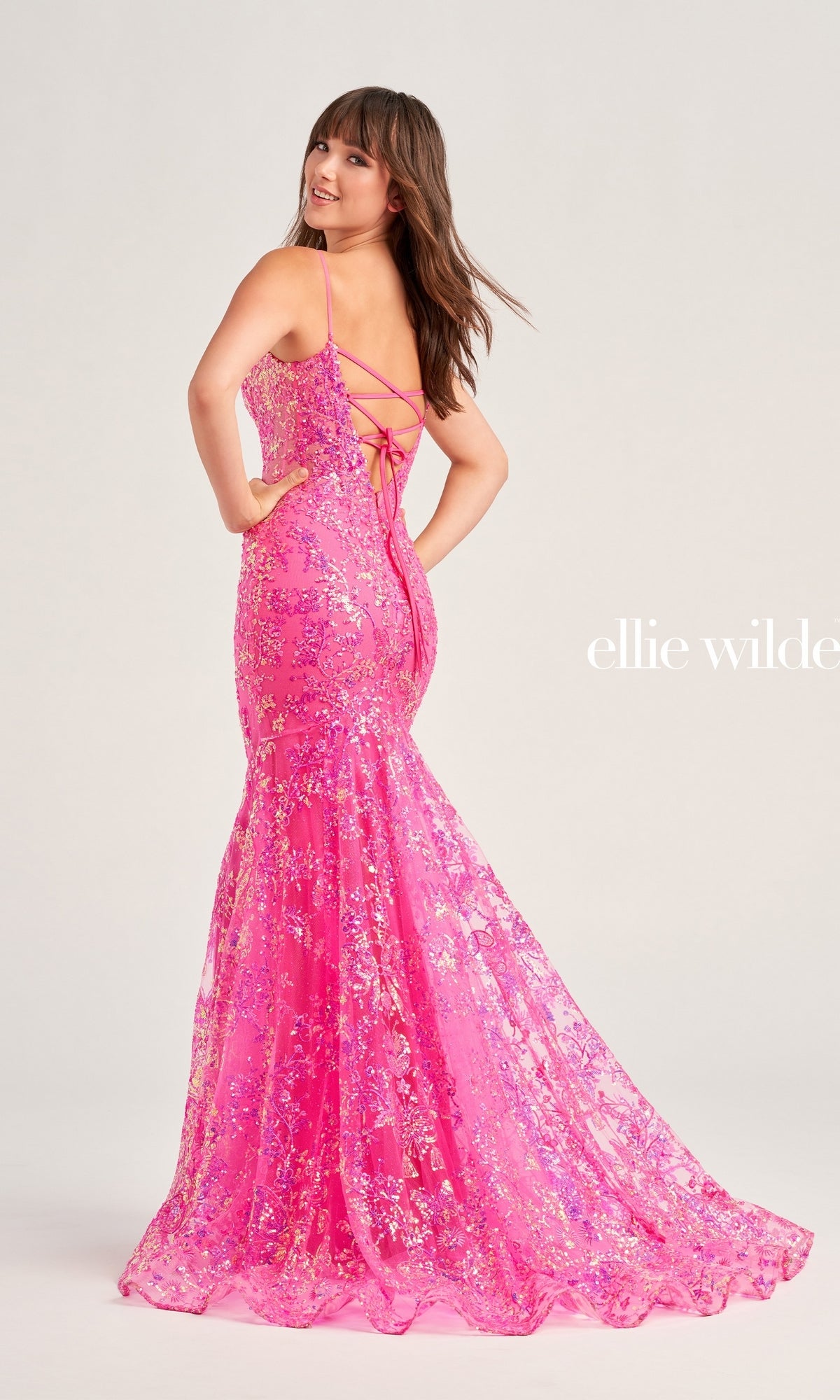 A woman models the Ellie Wilde Designer Glitter Prom Dress EW35013, a bright pink, floor-length, sequin mermaid gown with thin straps and a lace-up back, set against a plain background.