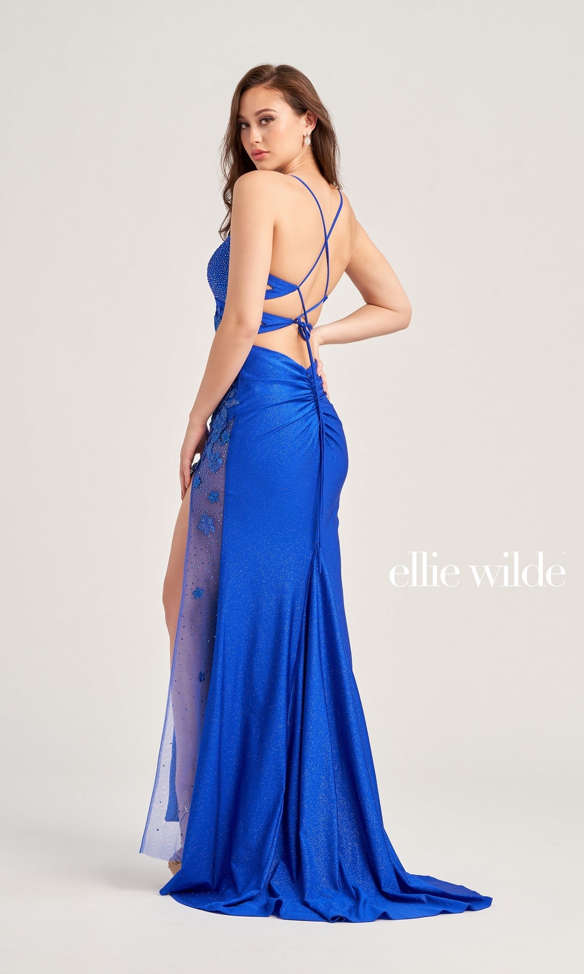 A woman wearing the Beaded-Lace Ellie Wilde Long Prom Dress EW35030, a bright blue floor-length gown with a high slit and crisscross open back, looks over her shoulder. The 