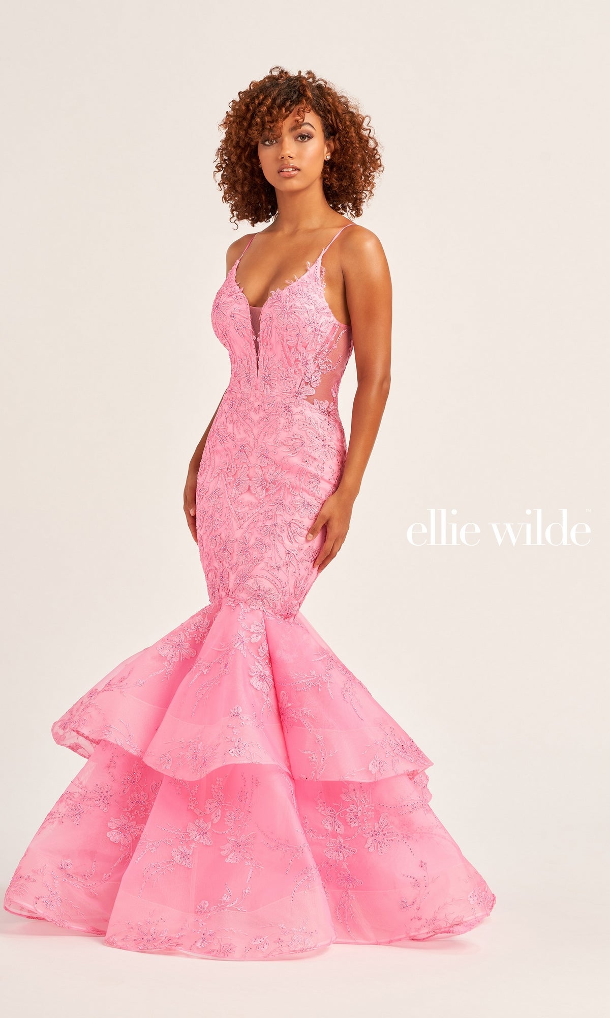 A woman poses against a plain background in the Ellie Wilde Long Lace Mermaid Prom Dress EW35038, a fitted pink mermaid gown with beaded lace details. The text 
