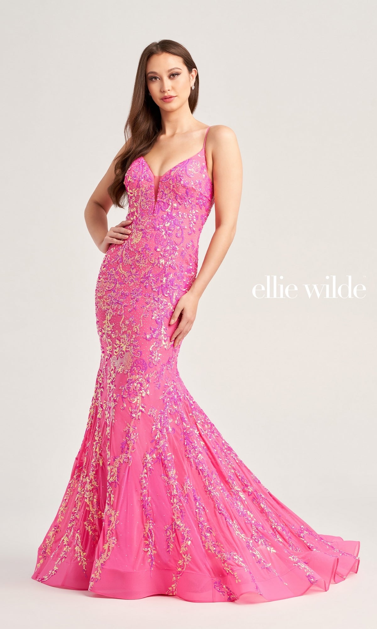 A woman wears the Ellie Wilde Long Mermaid Designer Dress EW35048—fitted, bright pink with sequin floral embellishments—standing against a plain background. 